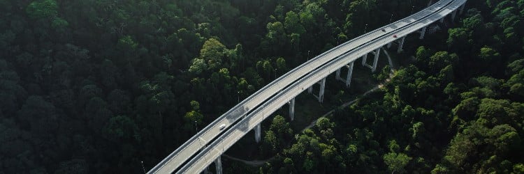 Aerial view of a winding highway cutting through dense green jungle, with vehicles traveling on the elevated roadway.